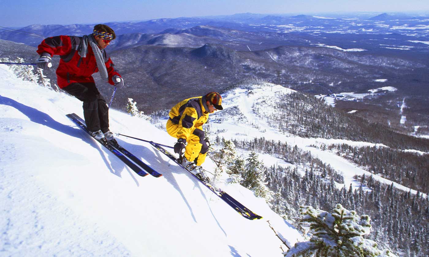 Two skiers skiing down snow capped mountainside on bright sunny day